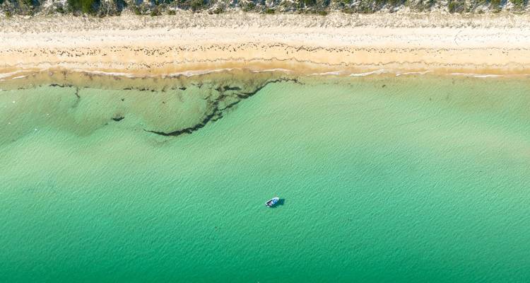 Prise de vue aérienne par drone d'un bateau solitaire flottant sur une eau d'un vert émeraude éclatant près d'un rivage de sable avec des motifs de marée texturés.