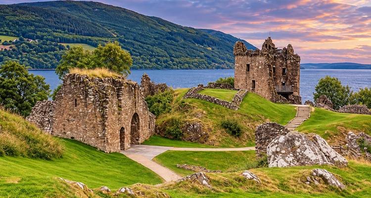 Les teintes chaudes du coucher de soleil illuminent les tours de pierre du château d'Urquhart avec le Loch Ness et les collines ondulantes au-delà.