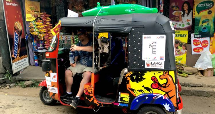 De deelnemer zit in een felgekleurd tuk-tuk met sponsorlogo's erop op een dorpsstraat.