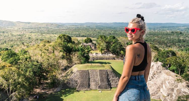 Un voyageur portant des lunettes de soleil rouges se tient au sommet d'une pyramide maya surplombant des ruines de jungle.