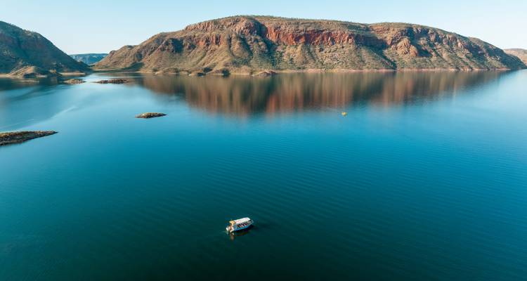 Vue aérienne d'un petit bateau de tourisme traversant les eaux turquoise calmes d'un vaste lac avec des falaises de roche rouge et des collines se reflétant à la surface.