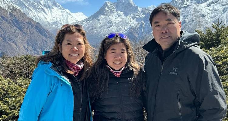 Familie van drie personen glimlacht voor het dramatische Himalaya-landschap met duidelijk zicht op sneeuwbedekte bergtoppen.