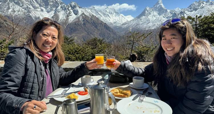 Twee vrouwen klinken met glazen sap terwijl ze buiten eten met een prachtig besneeuwd Himalaya-uitzicht op de achtergrond.