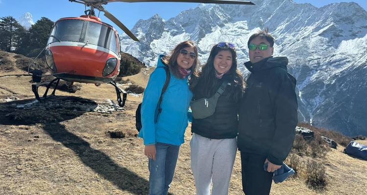 Drie trekkingsliefhebbers staan naast een rode helikopter op een grassy bergrug met torenhoge Everest-pieken die boven hen uitsteken.