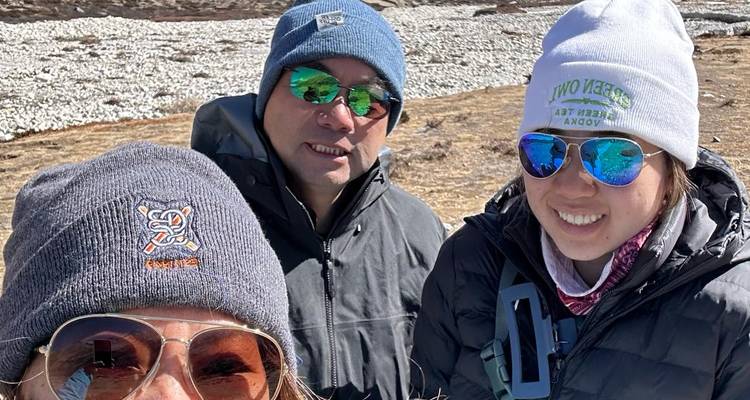Close-up selfie van drie trekkkers in winterkleding met een stenig rivierbedding en besneeuwde pieken op de achtergrond.