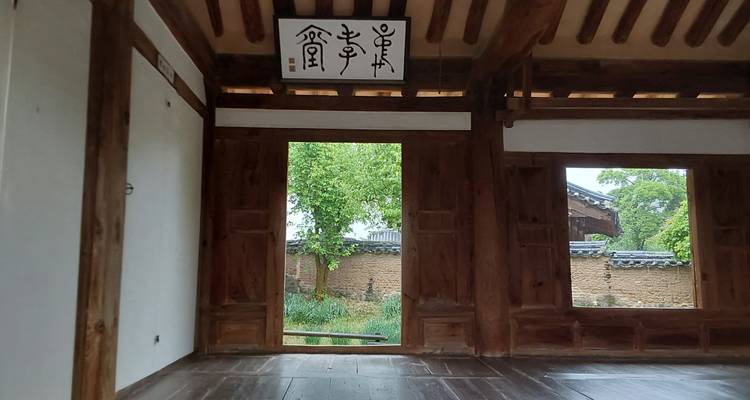 Intérieur en bois simple d'un bâtiment coréen traditionnel avec des portes ouvertes révélant une cour en terre battue et de la verdure à l'extérieur.