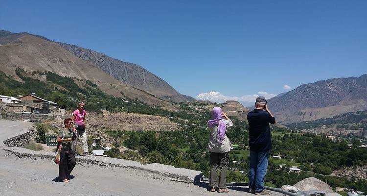 Travellers stop on a mountain roadside, photographing the wide green valley and distant snow peaks of northern Pakistan.
