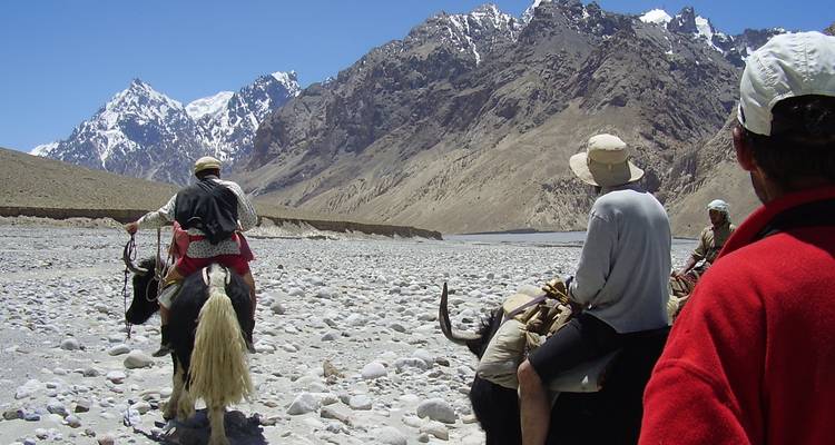 Travellers on yaks follow a rocky riverbed toward dramatic snow-capped Karakoram peaks under a clear blue sky.