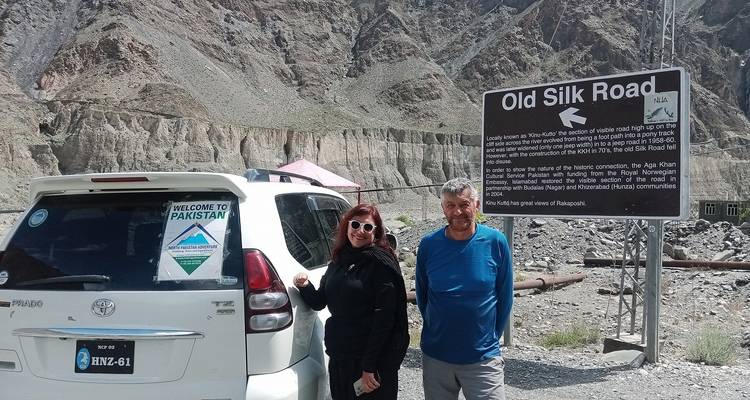 Two tourists stand beside a 4×4 and an 'Old Silk Road' sign against a cliff-lined valley.