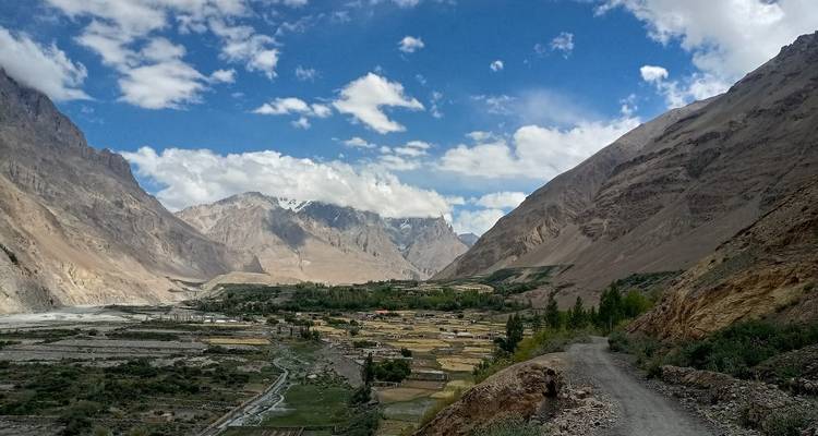 Panoramic view of a lush valley village framed by steep, rugged mountains and patchwork fields beneath blue skies.