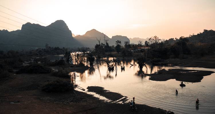 Lumière douce du coucher de soleil sur une rivière tranquille avec des villageois qui pataugent et des pics karstiques en silhouette dans le lointain.