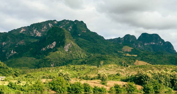 Chaîne de montagnes calcaires verdoyantes s'élevant au-dessus de terres basses forestières sous un ciel nuageux dans les zones rurales du Laos.