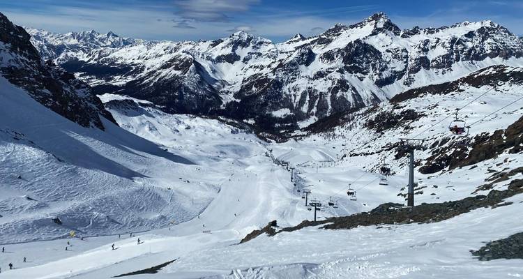 Breiter Panoramablick über ein verschneites Skigebiet mit von hohen gezackten Gipfeln und Sesselliften gesäumtem Tal.