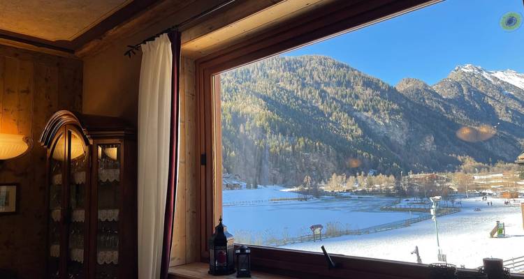 Großes Bildfenster mit Blick auf ein verschneites Tal, einen gefrorenen See und einen sonnigen blauen Himmel von einem warmen Innenraum aus.