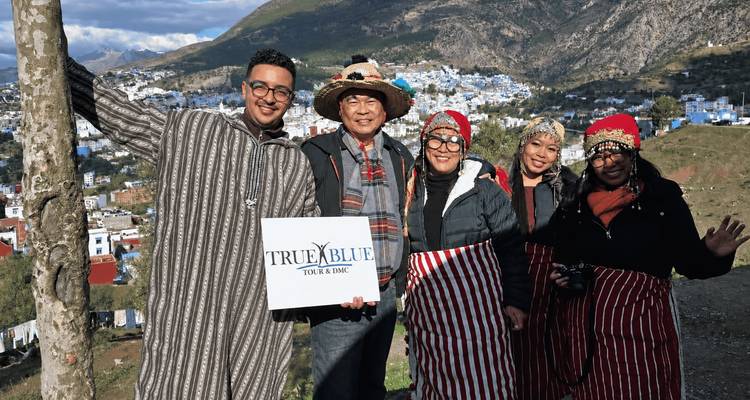 Un groupe de touristes en robes rayées pose avec un panneau de visite touristique surplombant le flanc de montagne bleu peint de Chefchaouen.