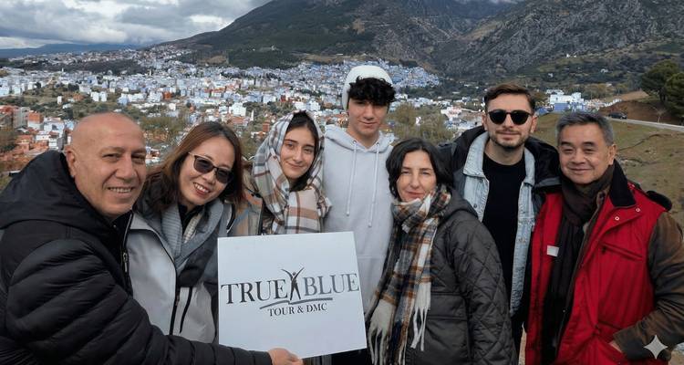 Groupe de voyageurs portant des écharpes et des vestes tenant une pancarte de visite touristique avec le paysage urbain bleu de Chefchaouen en arrière-plan.