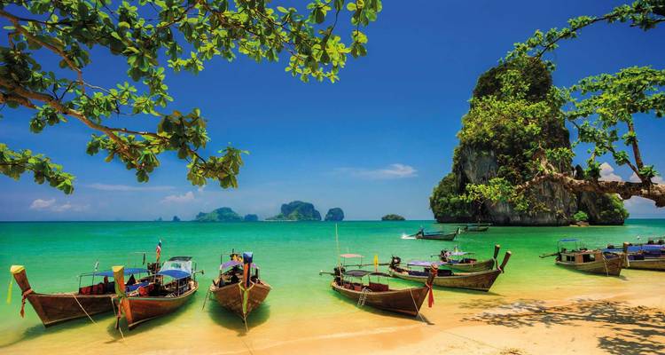 Plage tropicale avec des bateaux à longue queue sur une eau turquoise et des karsts calcaires sous un ciel bleu dégagé.