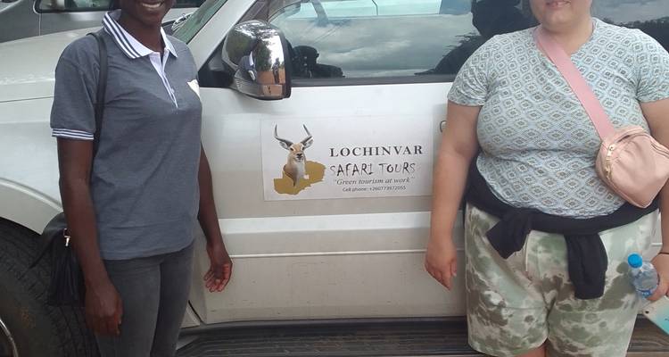 Two travellers stand beside a safari tour vehicle with company logo in a parking area.
