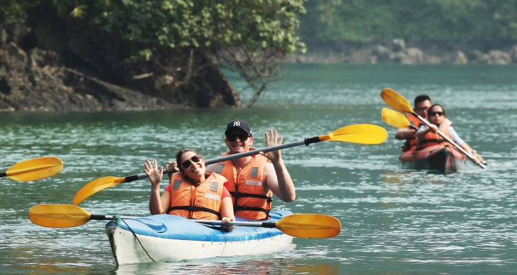 Des voyageurs heureux font du kayak dans les eaux turquoise d'une baie calcaire, saluant la caméra de la main.