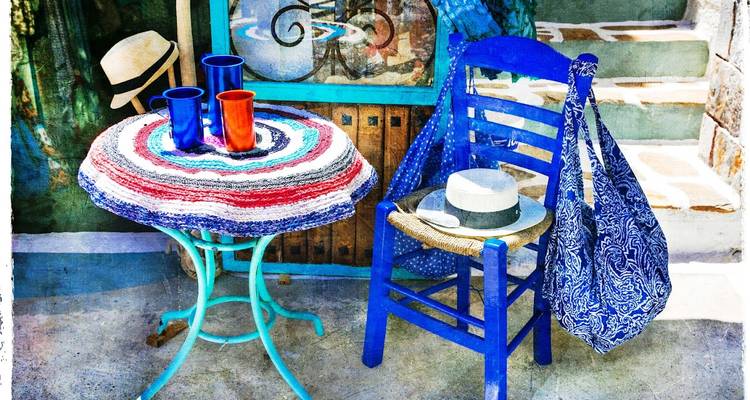 Artistic still life of colorful cups, woven tablecloth, blue chair and summer hat on Greek patio.
