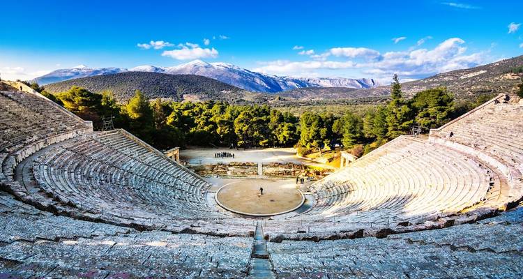 Ancient stone amphitheatre surrounded by forest with snow-capped mountains in distance.