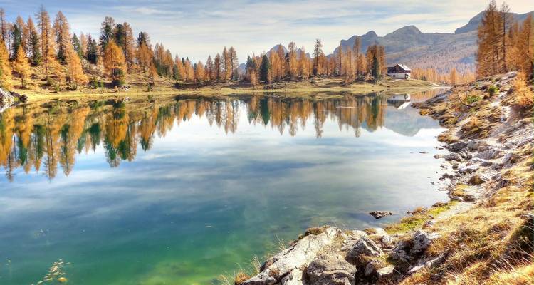 Un lago alpino cristalino refleja árboles de alerce dorado y lejanos picos rocosos junto a una cabaña de montaña solitaria.