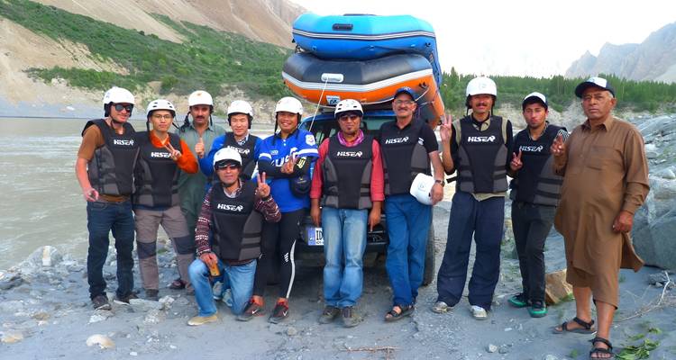 Rafting team wearing helmets and life vests pose beside an inflatable raft near a fast river.