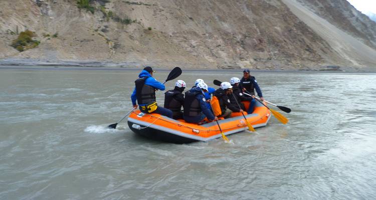 Adventurers paddle an orange inflatable raft down a wide mountain river surrounded by steep rocky slopes.