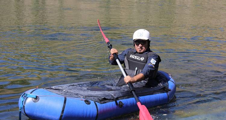 Kayaker paddles a small blue inflatable craft on calm alpine water wearing helmet and PFD.