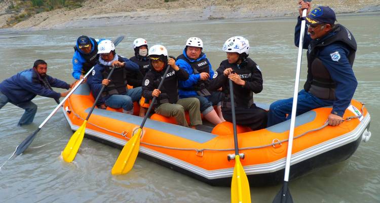 Team in helmets and jackets prepares an orange raft on a silty riverbank while a guide steadies it.