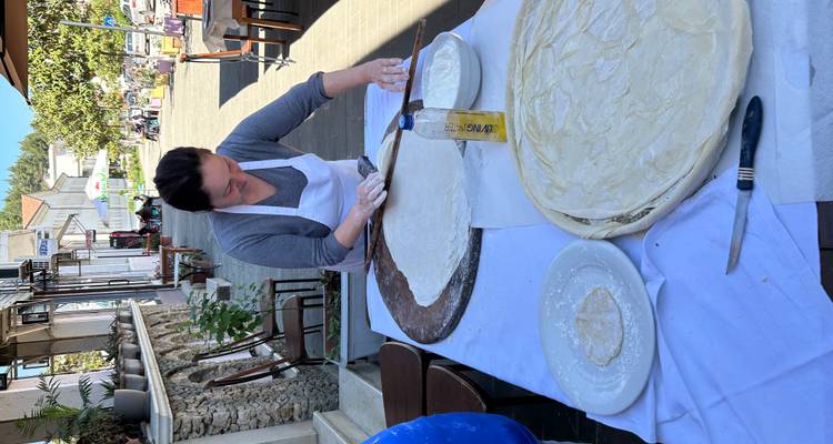 Een kok rolt grote cirkels deeg uit op een tafel buiten een dorpsrestaurant op een zonnig straatje.
