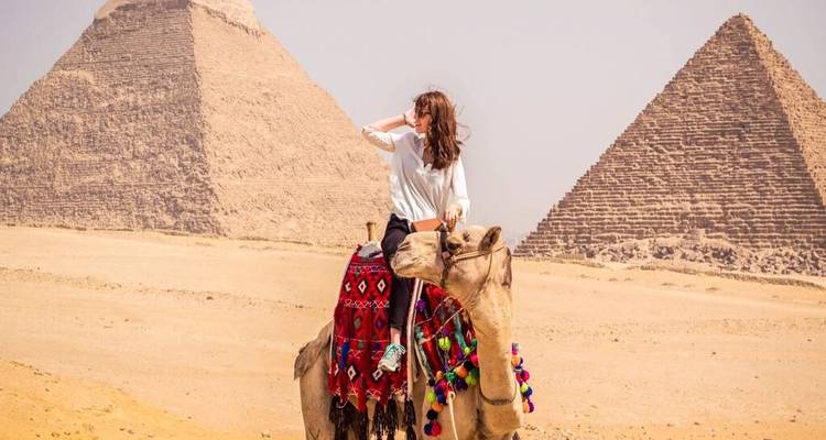 Woman riding decorated camel in front of pyramids on a hazy desert day.