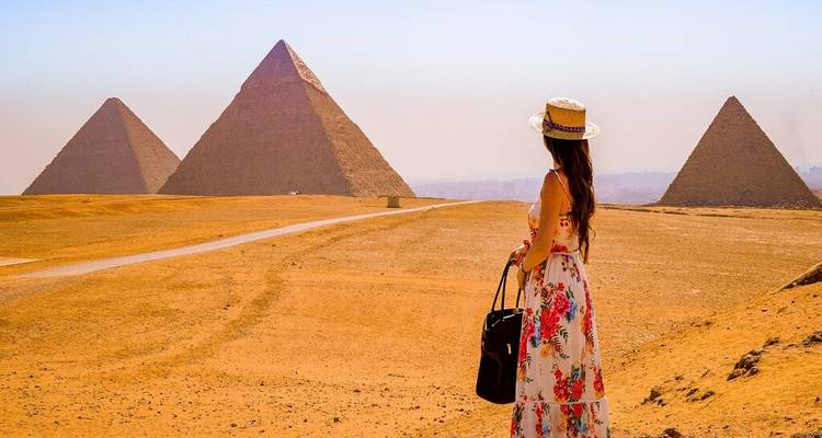 Woman in floral dress with tote bag looking toward four pyramids across sandy plateau.