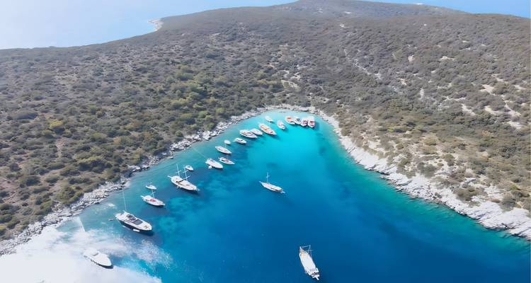 Drone-opname van zeilboten voor anker in een smalle, levendig blauwe inham omgeven door kale heuvels.