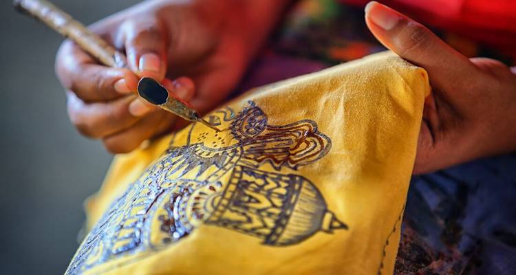 Gros plan sur des mains peignant un motif complexe sur un tissu jaune à l'aide d'un fin pinceau dans un atelier de batik.