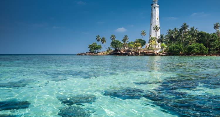 Crystal-clear turquoise water surrounds a small rocky island topped with a white lighthouse and palm trees.