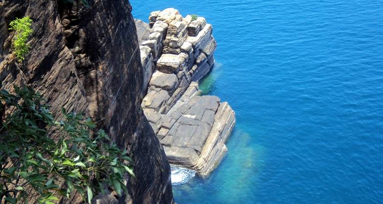 Steep rocky cliff plunges into vivid blue ocean water with visible underwater hues near the coast.