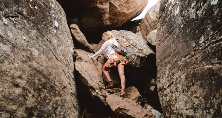A young woman climbs between large weathered boulders in a rocky canyon.