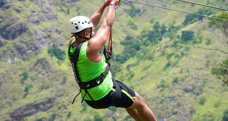 A man in bright green gear glides on a zip-line across a green mountain slope.