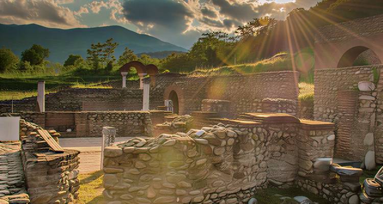 Sunbeams stream over ancient stone ruins set against forested hills.