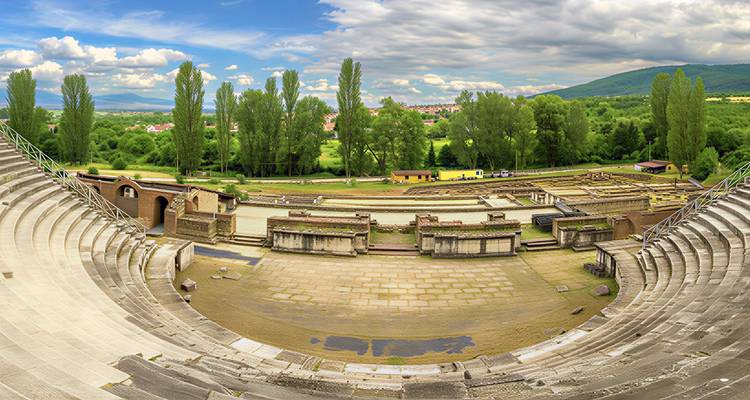 Panoramic view of a restored Roman amphitheatre surrounded by lush green trees.