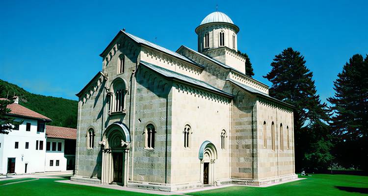 Stone Decani Monastery church stands on lush green lawn under clear blue sky.