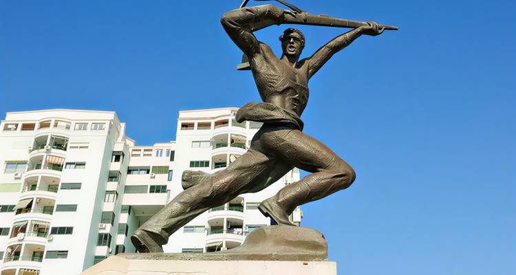 Bronze statue of a heroic figure holding a rifle aloft in front of modern apartment buildings against a clear blue sky.