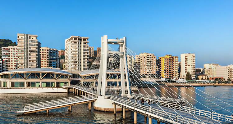 Modern cable-stayed pedestrian bridge leading to waterfront high-rise buildings over calm blue water.