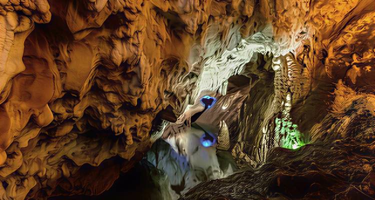 Colorfully lit cave interior with dramatic stalactites and stalagmites creating abstract rock patterns.