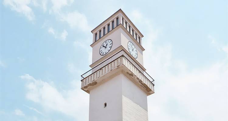 Pale pink clock tower with twin faces rising into a bright blue sky with light clouds.