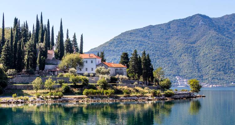 Monastère en pierre et grands cyprès se reflétant dans les eaux bleues et calmes de la baie de Kotor.