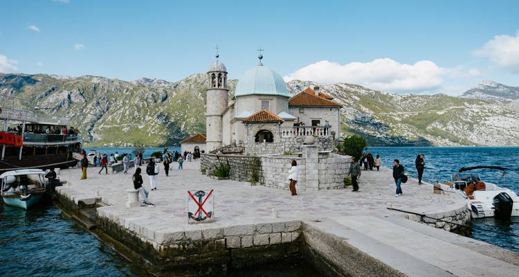 Steinige Inselkirche Unsere Liebe Frau der Felsen mit Besuchern, vor einer bergigen Küstenlinie und blauem Meer