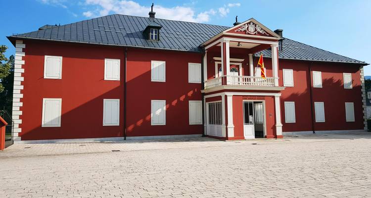Neoklassischer rot angestrichener Palast mit Balkon und Fahnenstangen auf einem gepflasterten Hof unter blauem Himmel