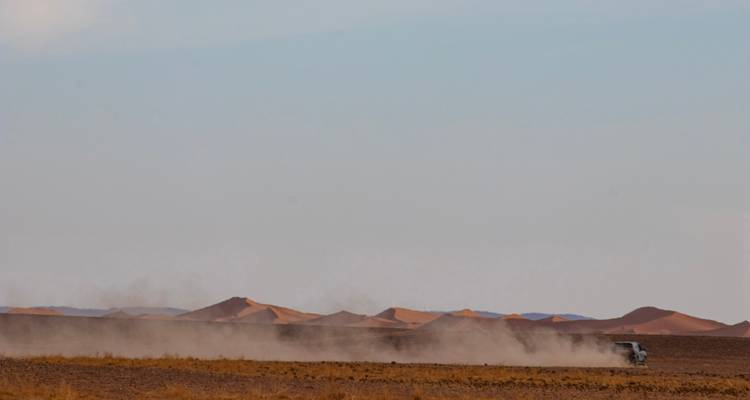 Distant 4x4 vehicle raises dust across a vast flat desert plain with dune ridge on horizon.